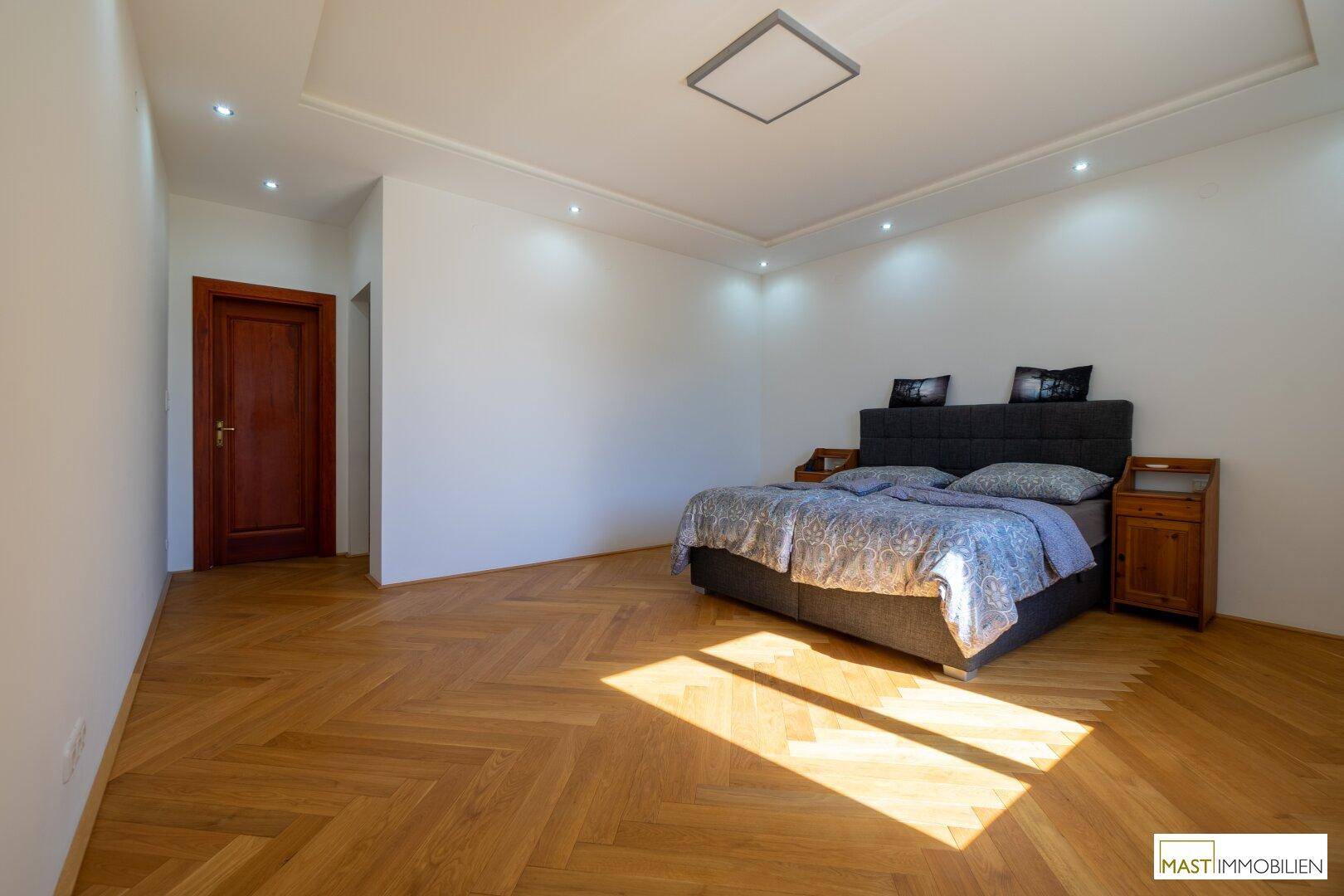 Helles Schlafzimmer mit Fischgrätparkett, Doppelbett und indirekter Deckenbeleuchtung.