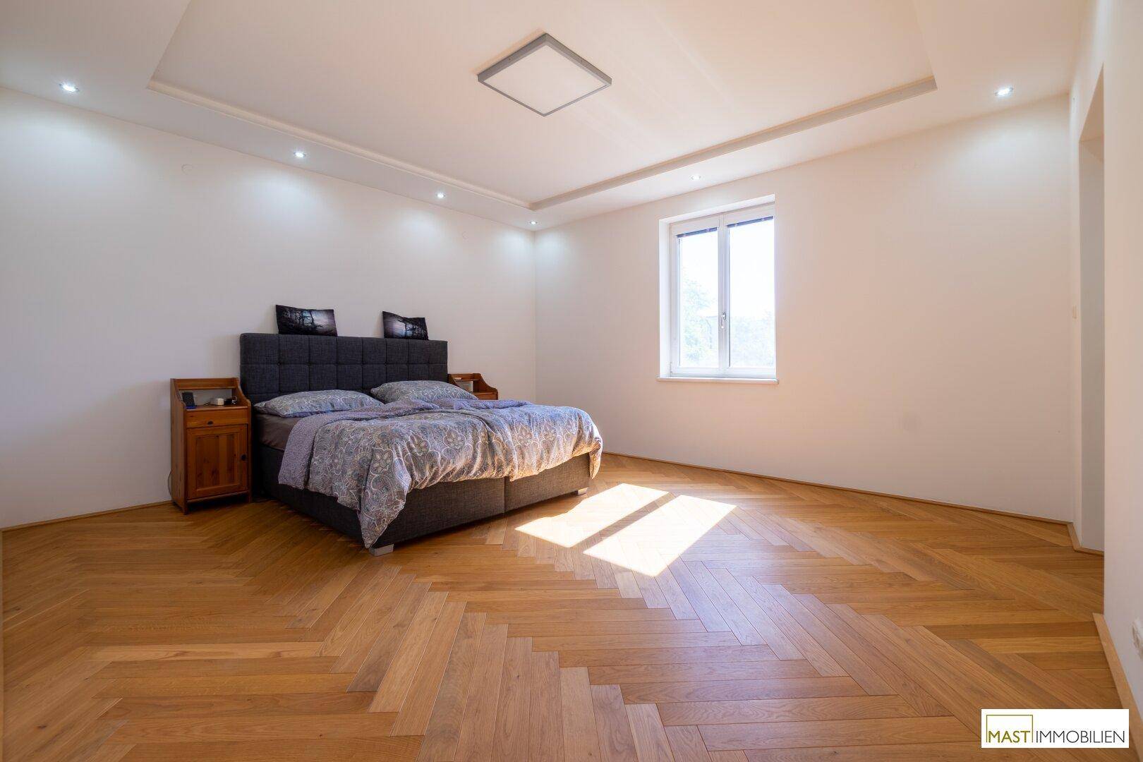 Geräumiges Schlafzimmer mit Fischgrätparkett, großem Fenster und viel Tageslicht.