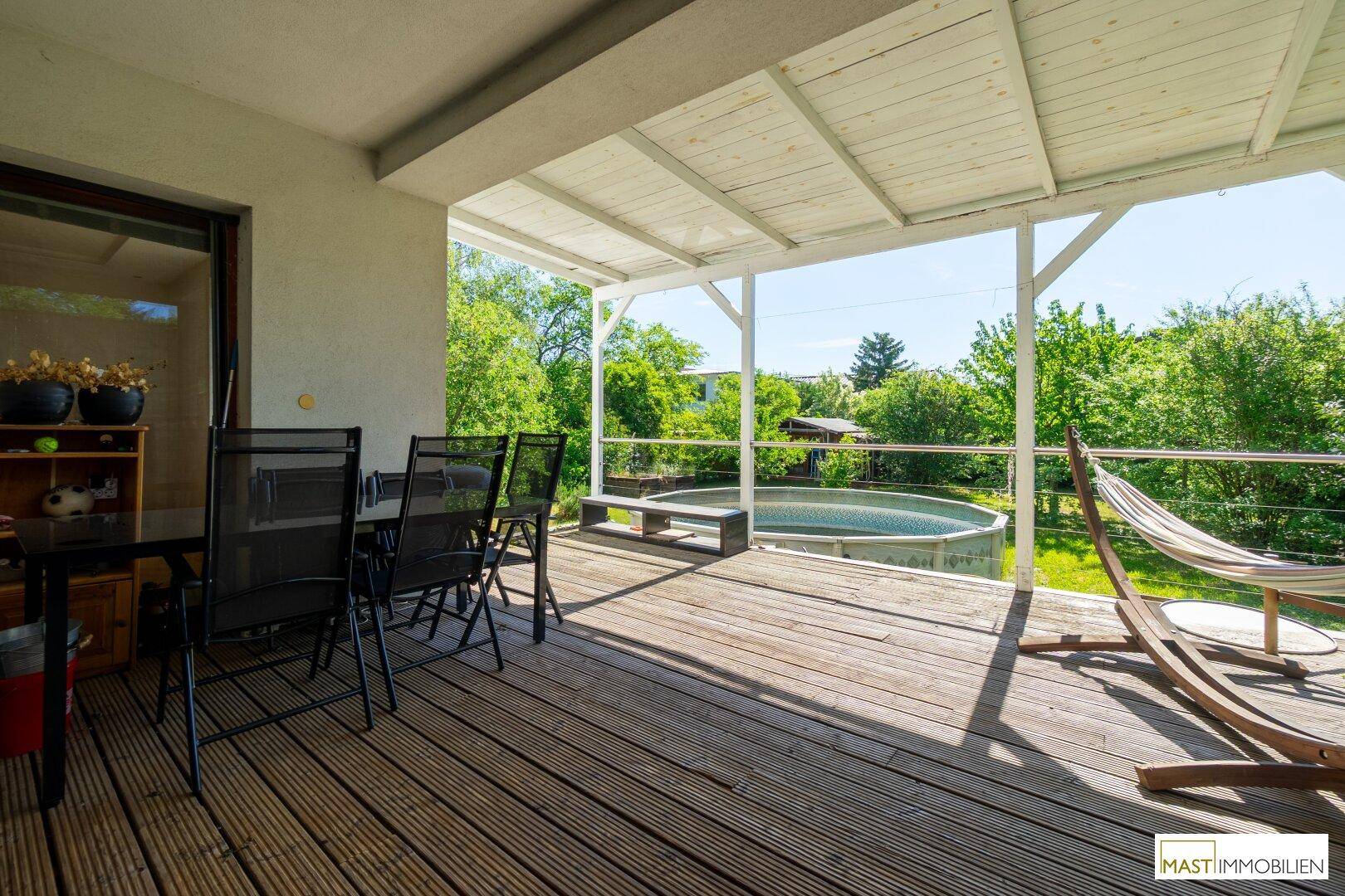 Überdachte Holzterrasse mit Sitzgelegenheiten, Hängematte und Blick auf den Garten mit Pool.