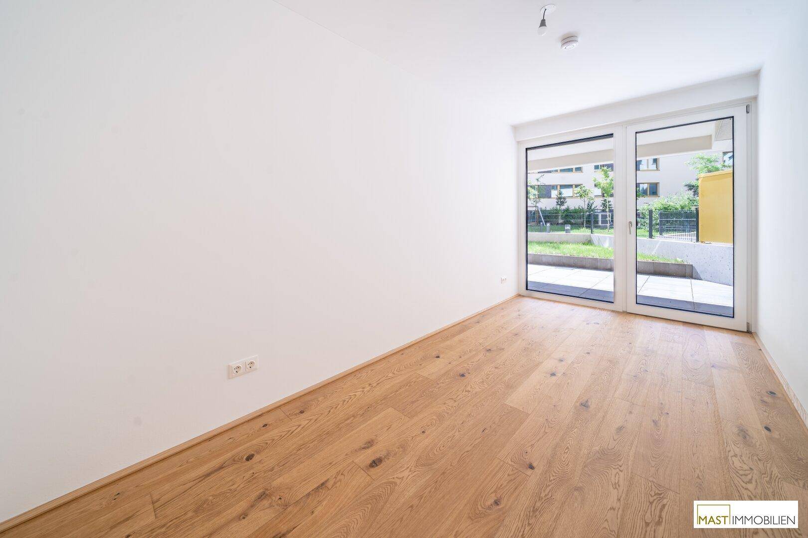 Leeres Schlafzimmer mit Holzboden und großen Fenstern zum Außenbereich.