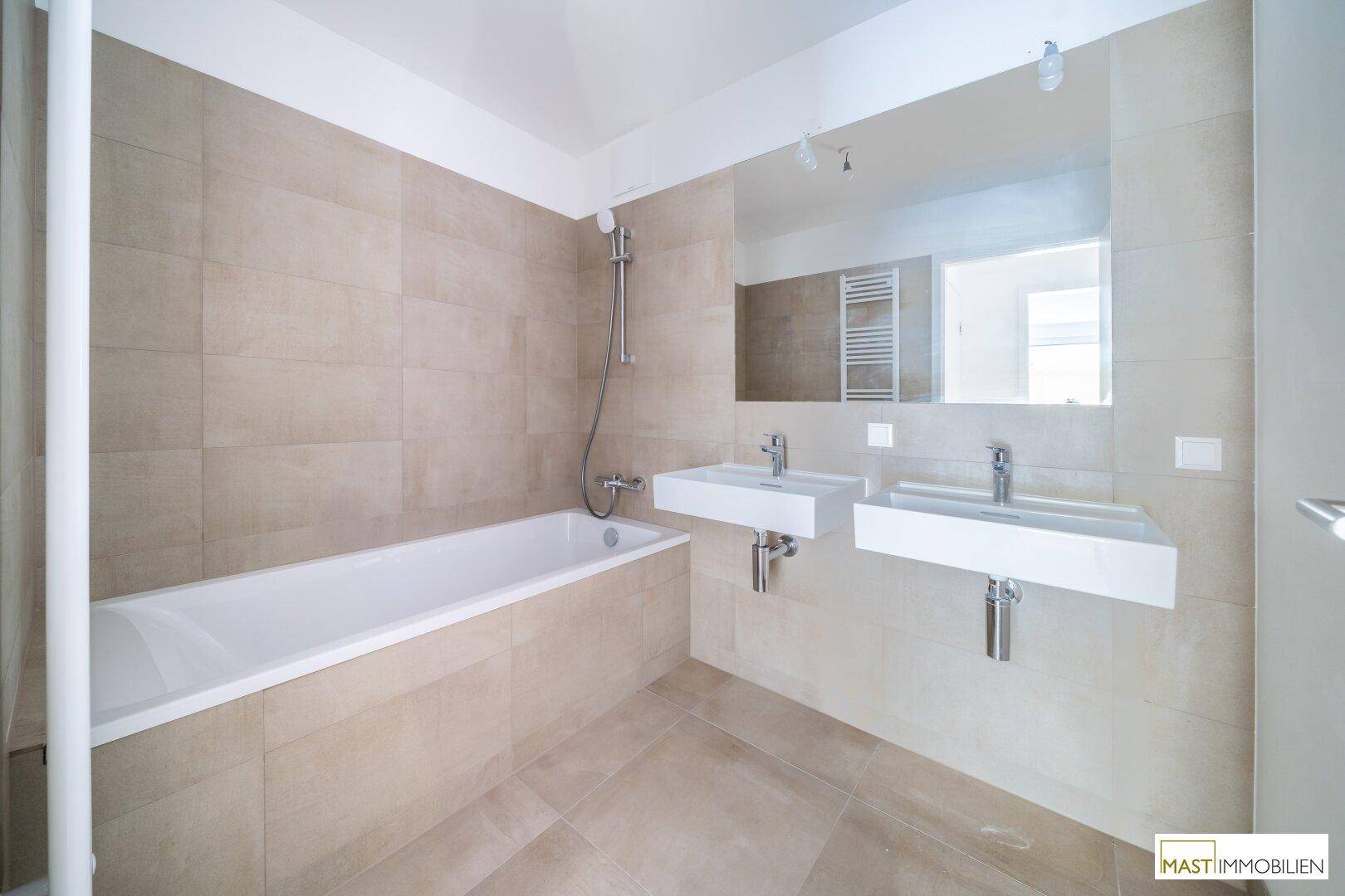 Helles Badezimmer mit beigefarbenen Fliesen, Badewanne mit Duschfunktion und Doppelwaschbecken.