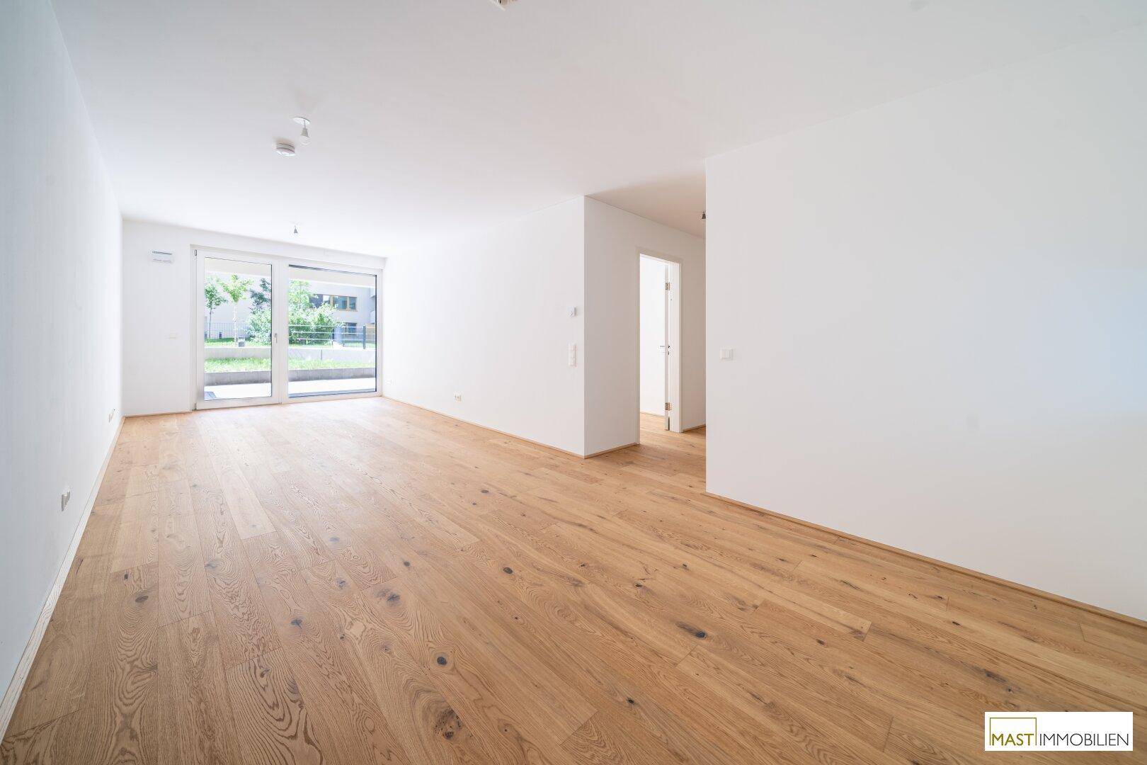 Heller, leerer Wohnraum mit Holzboden und Zugang zu einer Außenterrasse.