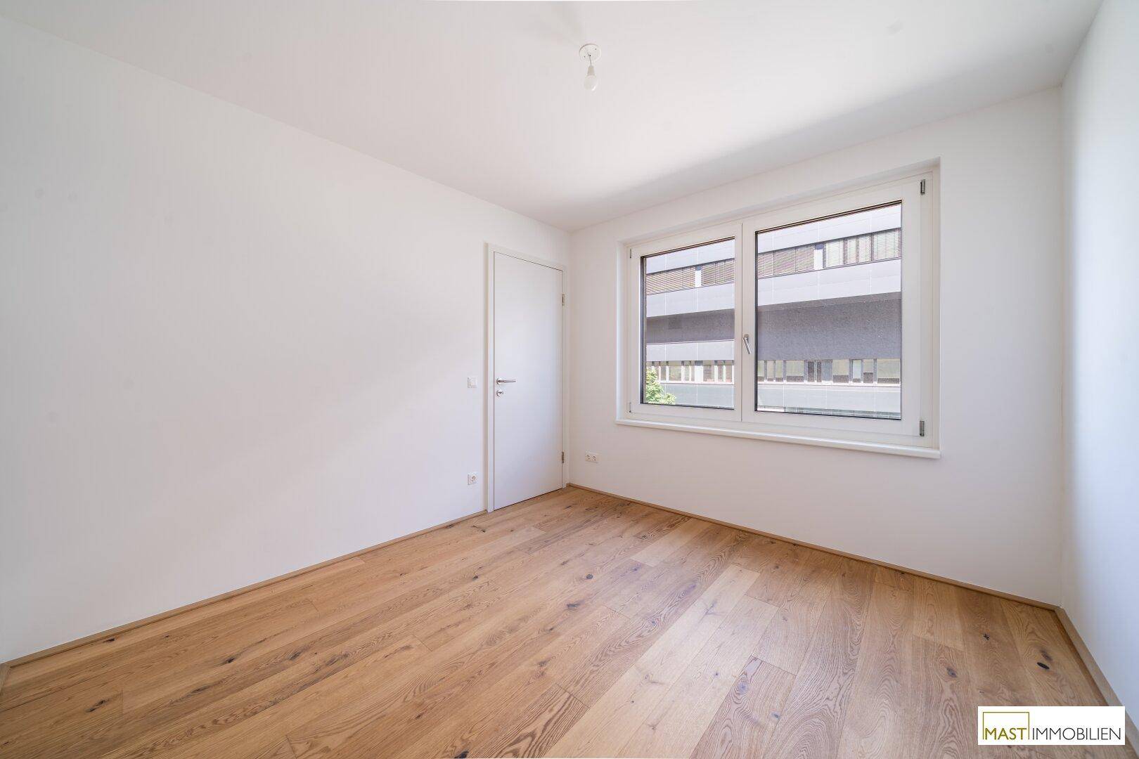 Geräumiger, heller Raum mit Holzparkett und einem großen Fenster, ideal für Schlaf- oder Arbeitszimmer.