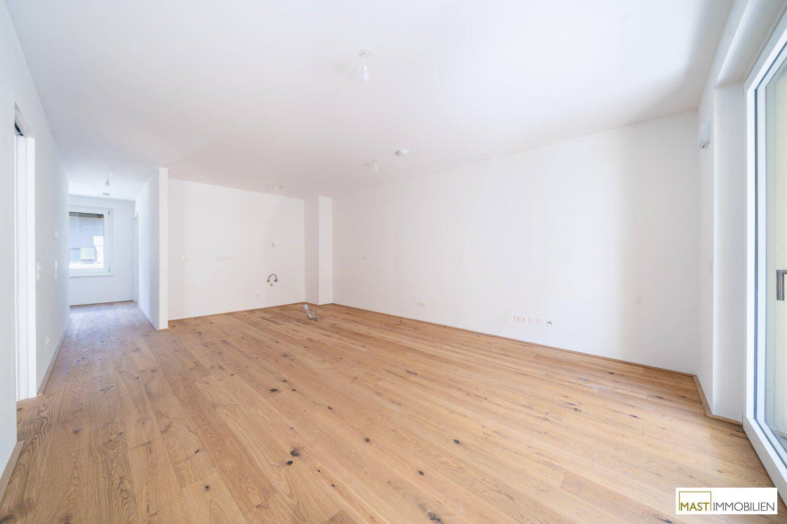 Großer, offener Wohnbereich mit Holzparkett und vorbereiteten Anschlüssen für eine Küche.