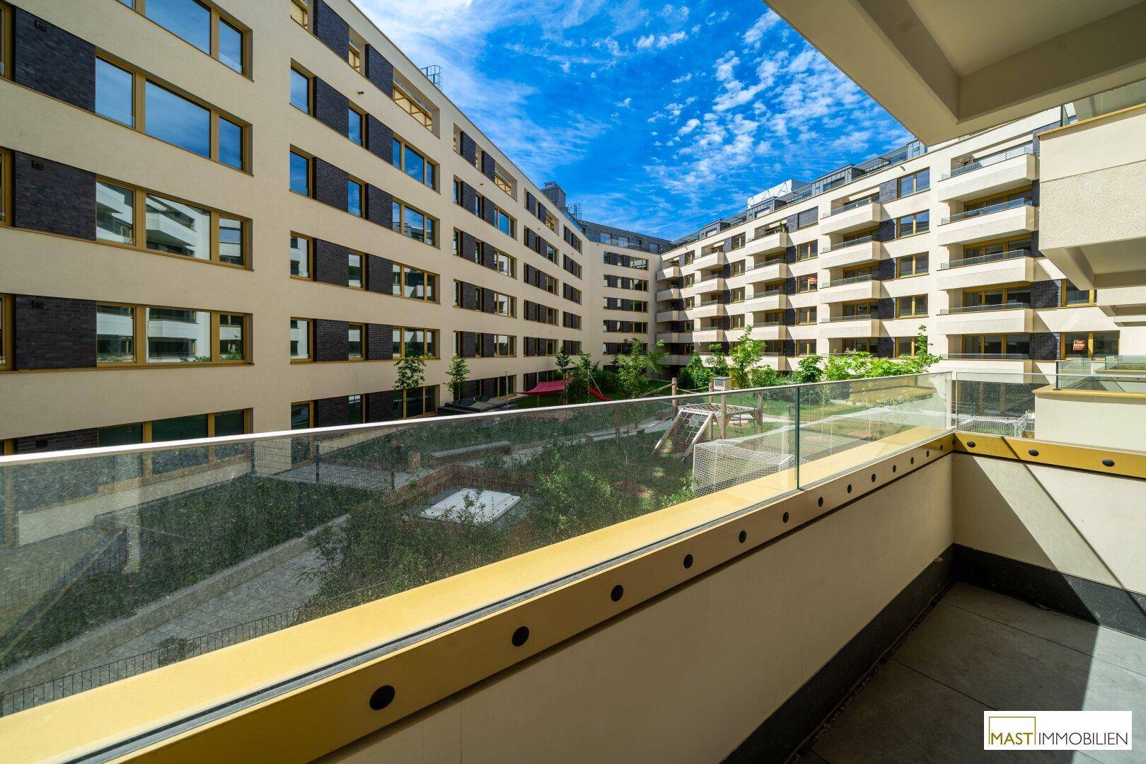 Balkon mit Glasgeländer und Fliesenboden, bietet einen weiten Blick auf den Innenhof und Spielplatz.
