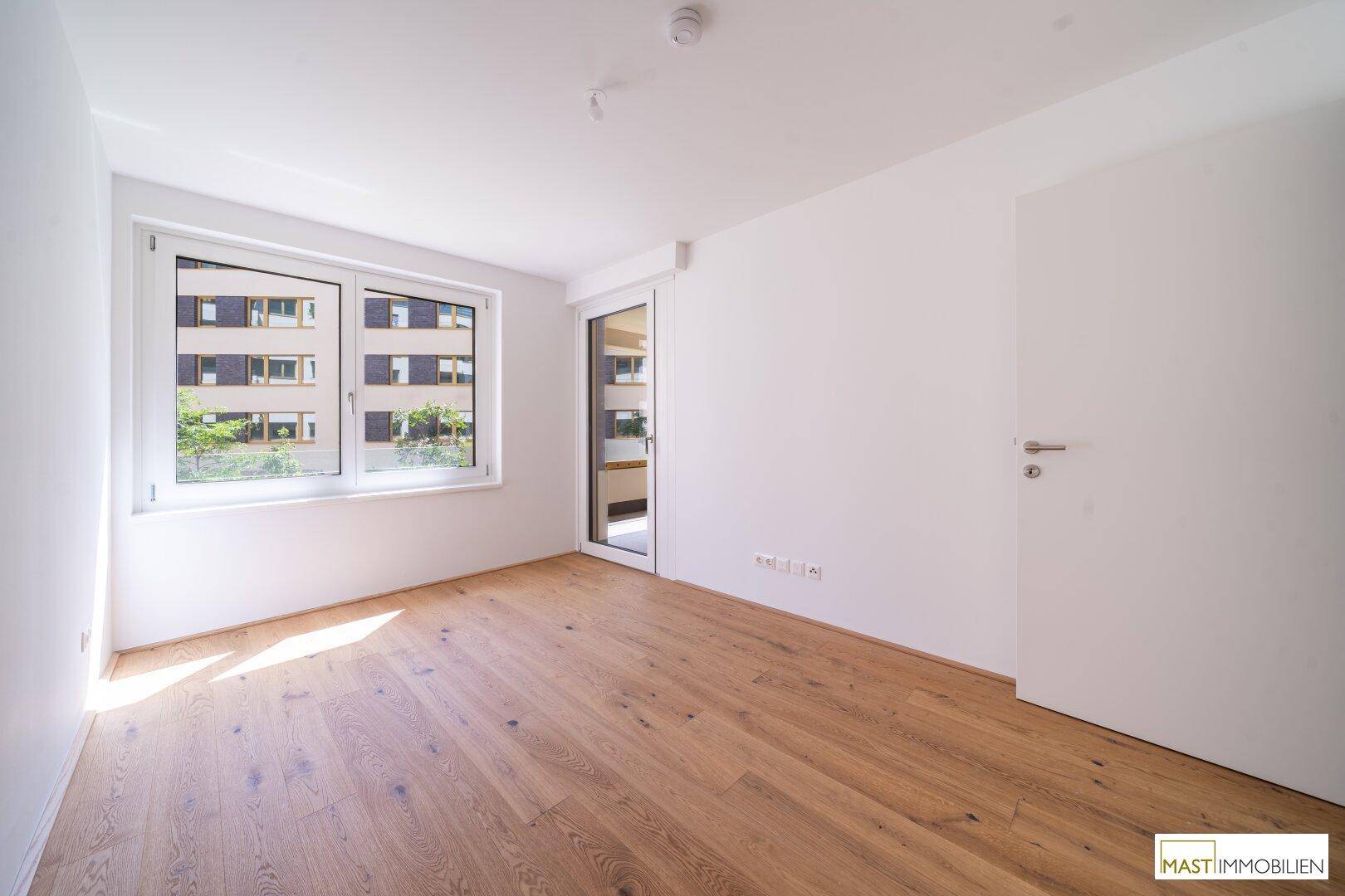 Helles Schlafzimmer mit Parkettboden und Fenster, das einen Blick auf die umliegenden Gebäude bietet.