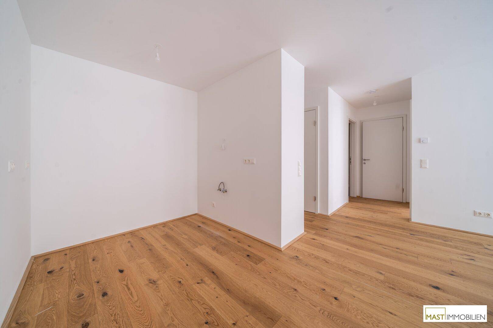 Leerer Raum mit Holzboden und Anschlüssen, möglicherweise für eine Küche oder Waschküche.