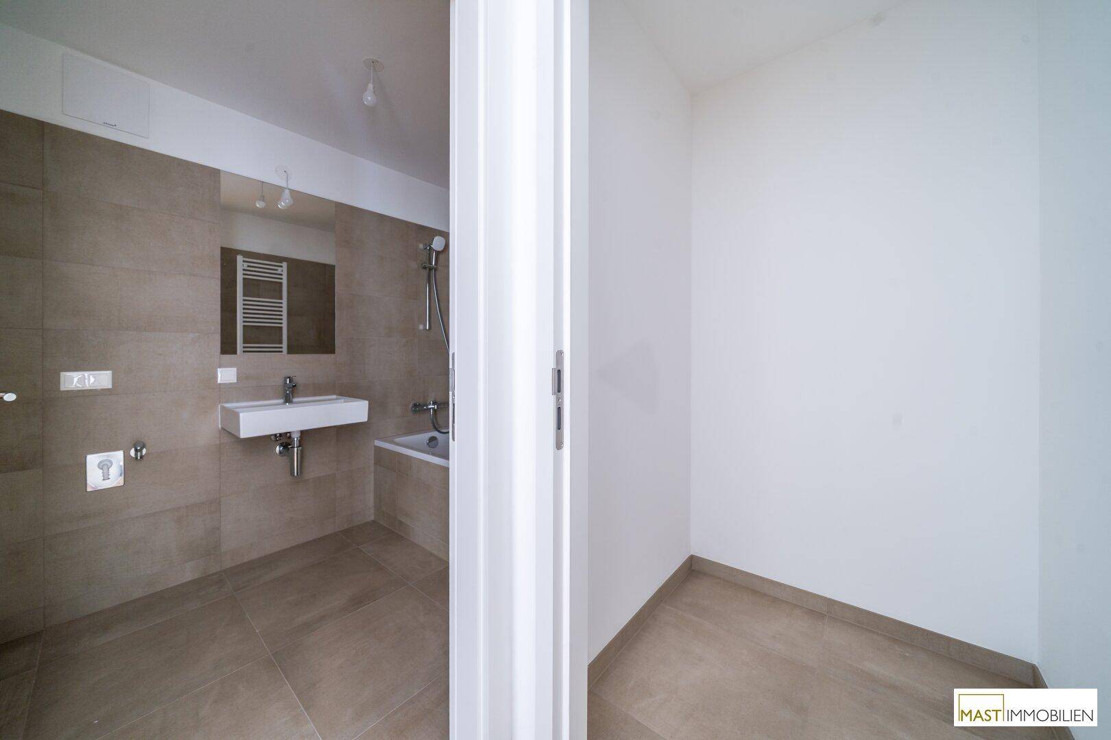 Blick durch eine Tür in ein modernes Badezimmer mit beigefarbenen Fliesen und Badewanne.