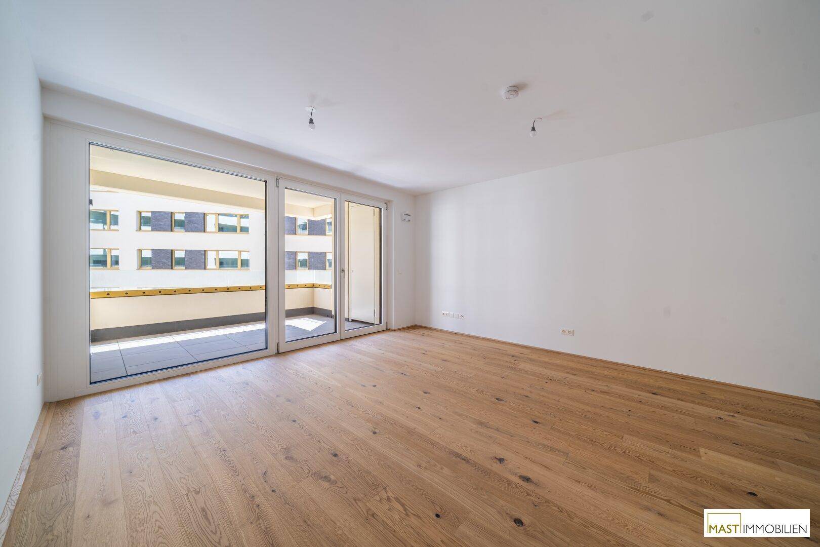 Lichtdurchfluteter Wohnraum mit hellem Holzboden und breiter Glasfront zum Balkon, bietet viel Tageslicht.