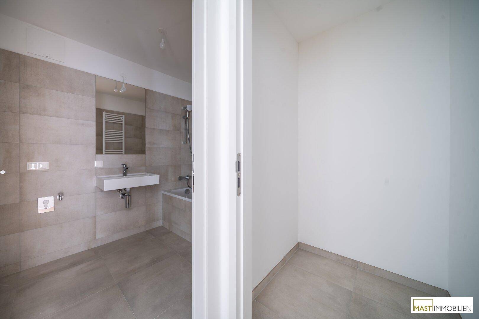 Modernes Badezimmer mit hellen Fliesen, Waschbecken und Badewanne, teilweise durch Türrahmen verdeckt.