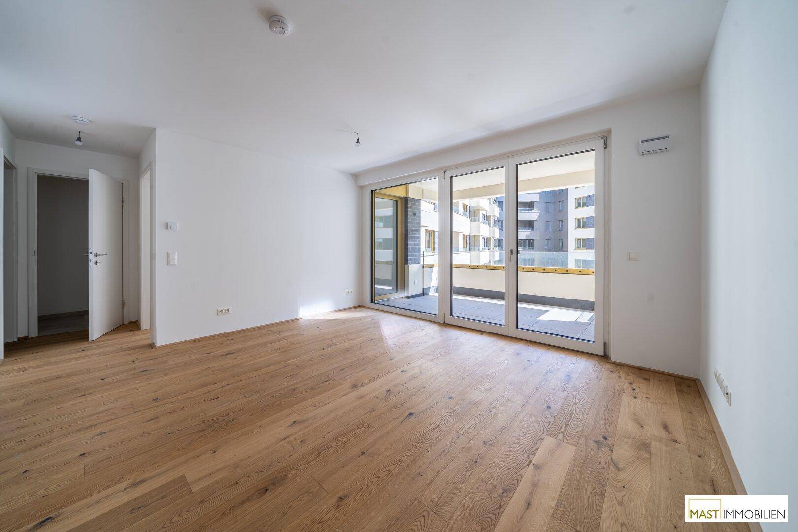 Helles, leeres Wohnzimmer mit Eichenparkettboden und Zugang zu einem großen Balkon.