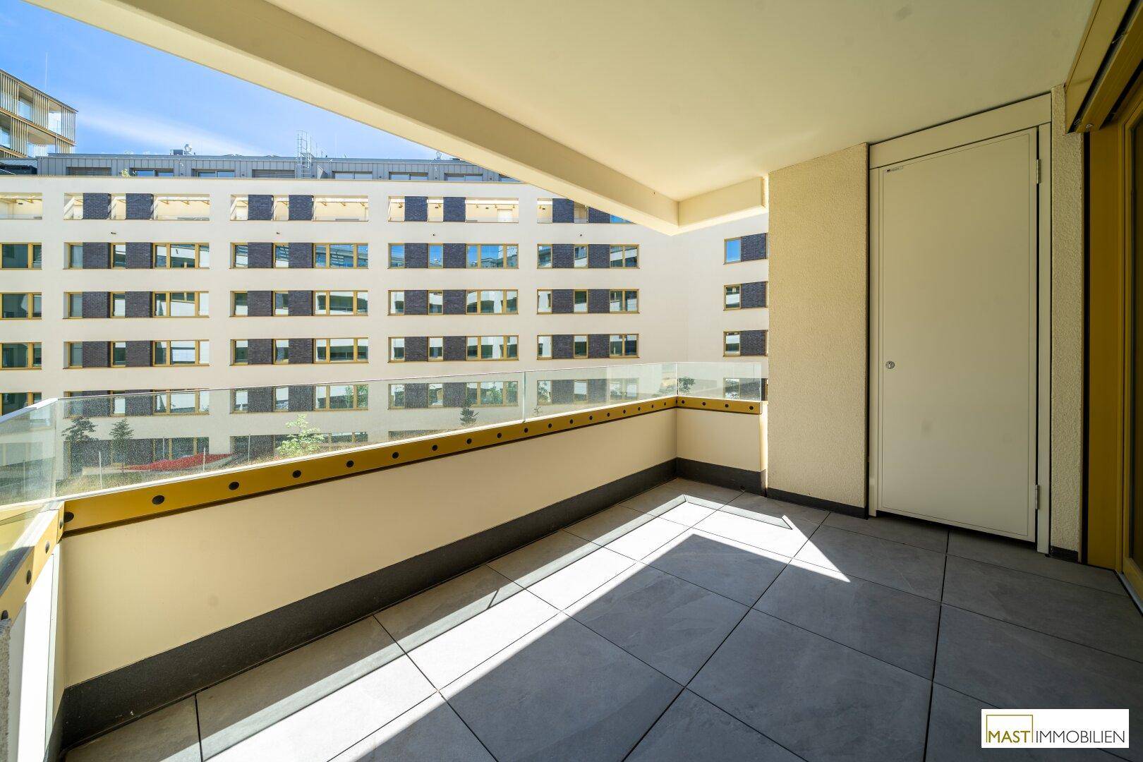 Sonniger Balkon mit Fliesenboden und Glasbrüstung, bietet einen weiten Blick auf die umliegende Bebauung.