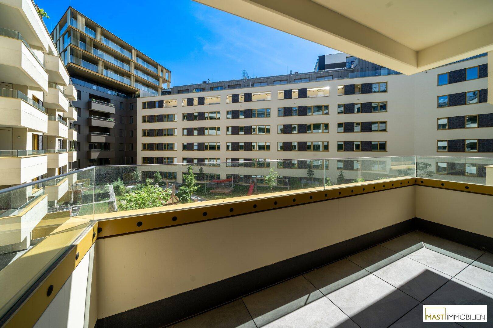 Großer Balkon mit Glasgeländer und Blick auf den begrünten Innenhof und die umliegenden Gebäude.