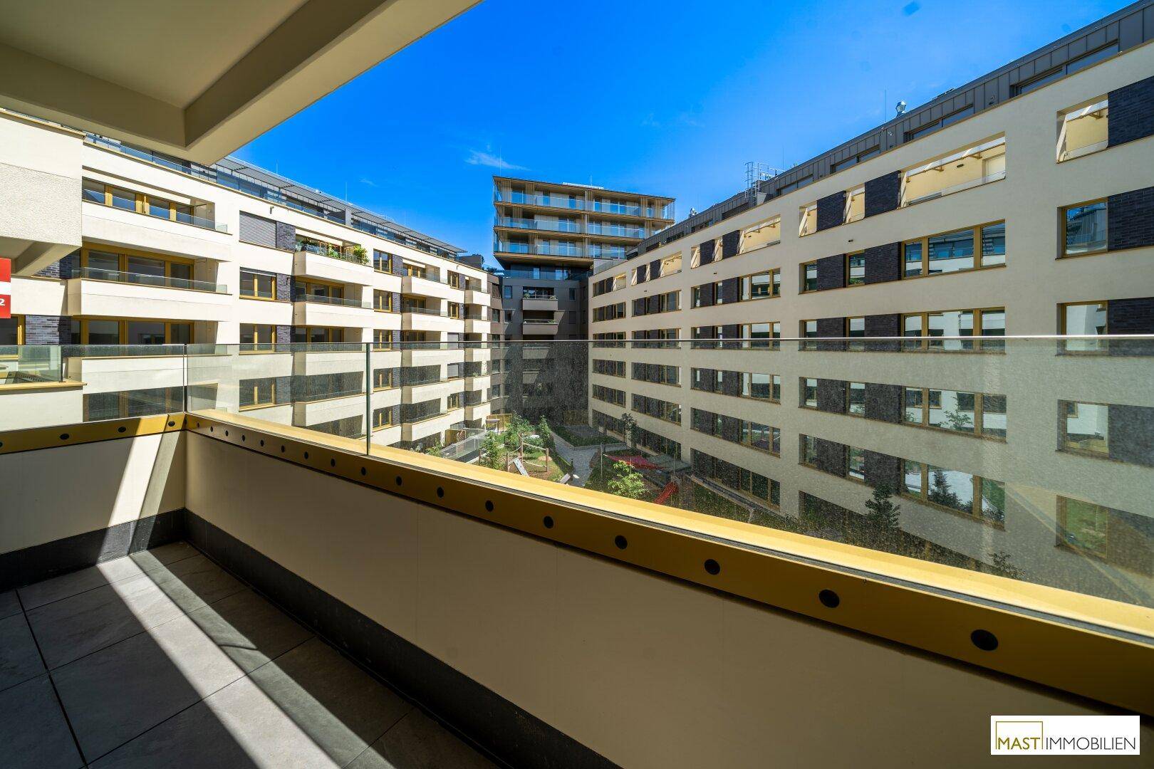 Balkon mit Glasgeländer und weitem Blick auf die modernen Gebäude und den grünen Innenhof.