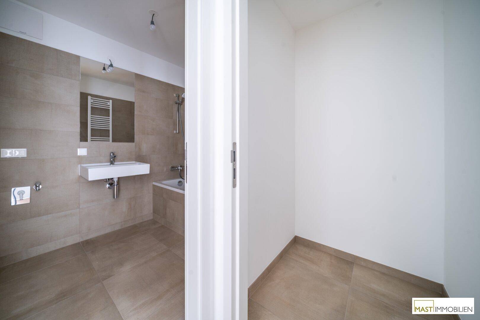 Blick in ein modernes Badezimmer mit Badewanne und Waschtisch, angrenzend an einen leeren Raum.