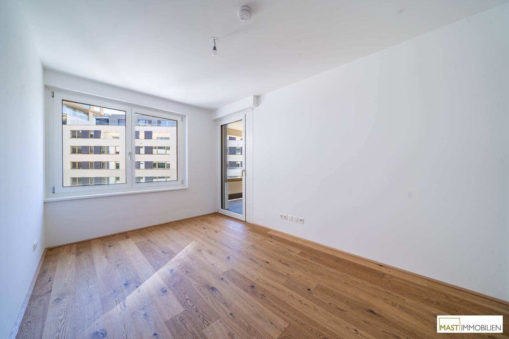 Helles Schlafzimmer mit Holzboden und großem Fenster, das viel Tageslicht hereinlässt.