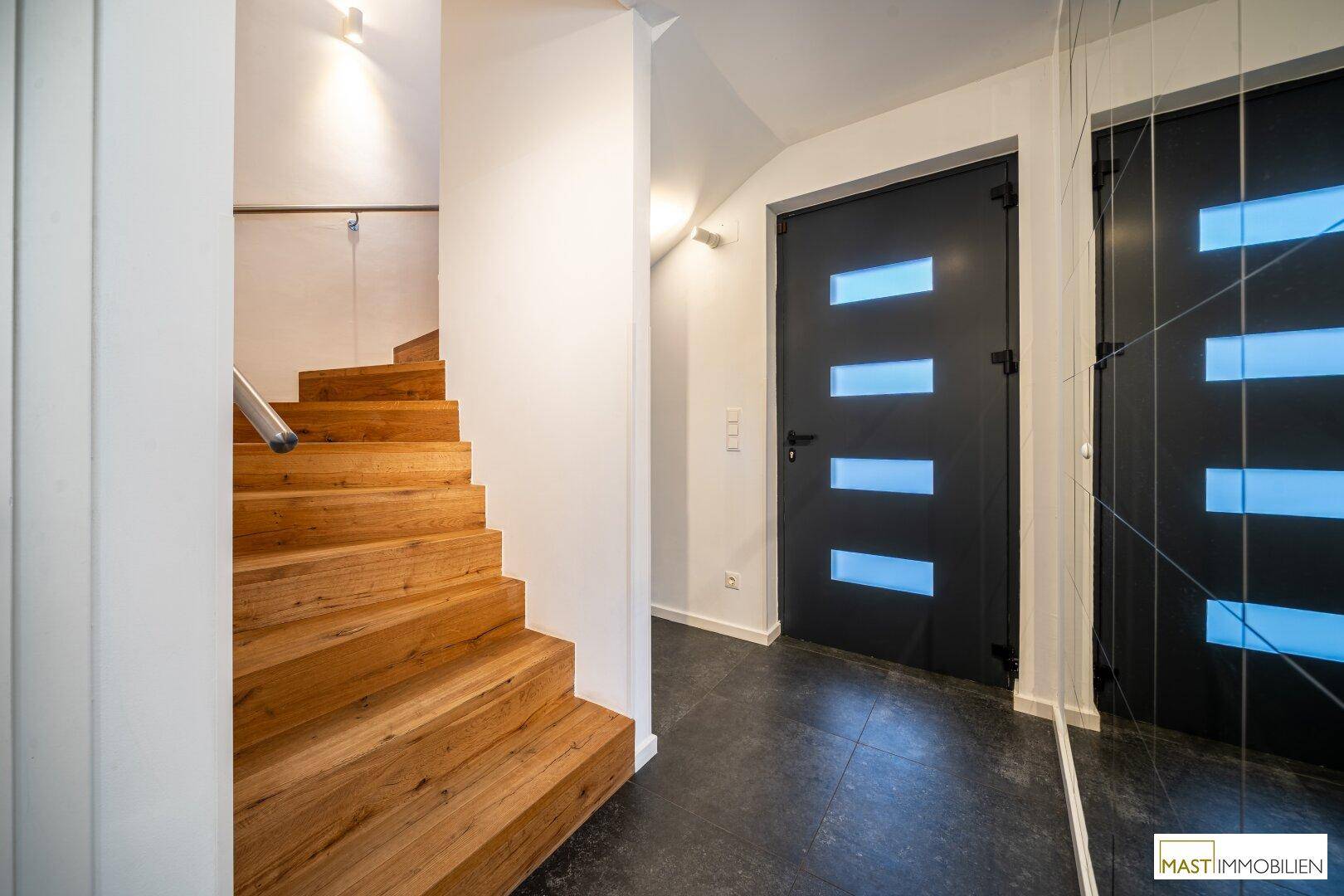 Heller Flur mit Holztreppe, moderner Eingangstür und einer dekorativen Spiegelwand.