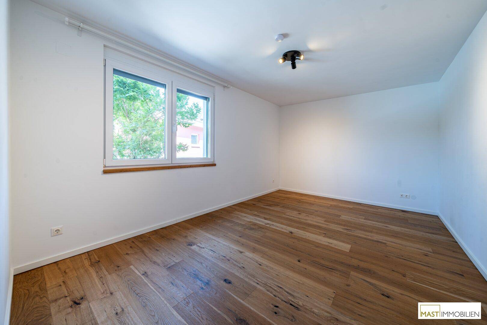 Helles Schlafzimmer mit Holzparkettboden und einem Fenster mit Blick ins Grüne.
