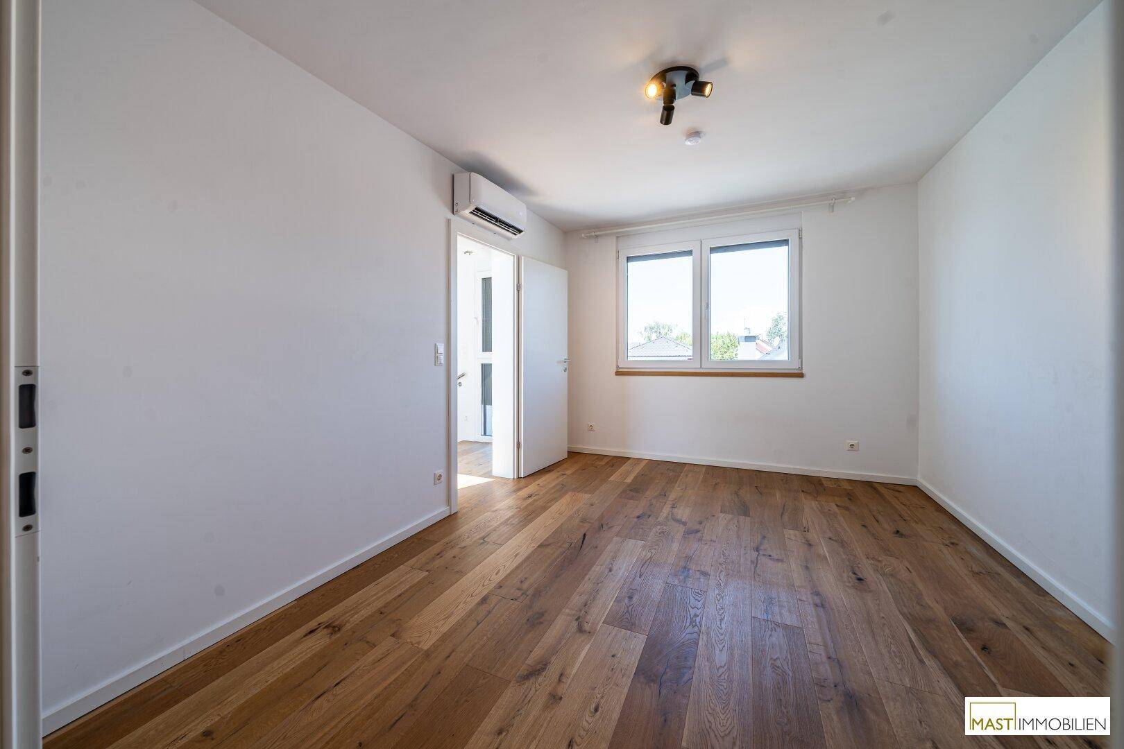 Geräumiges Schlafzimmer mit Holzparkett, Fenster und installierter Klimaanlage für angenehmes Klima.