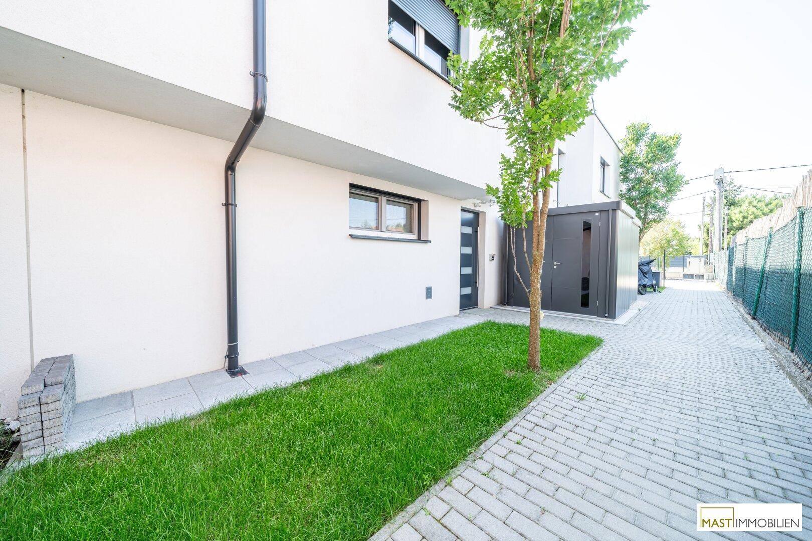 Seitenansicht des Hauses mit Rasenfläche, gepflastertem Weg und einem modernen Schuppen oder Carport.