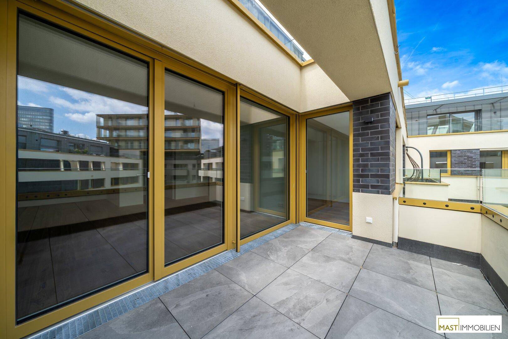 Heller Balkon mit großen Glasfronten und Blick auf die moderne Wohnanlage unter blauem Himmel.