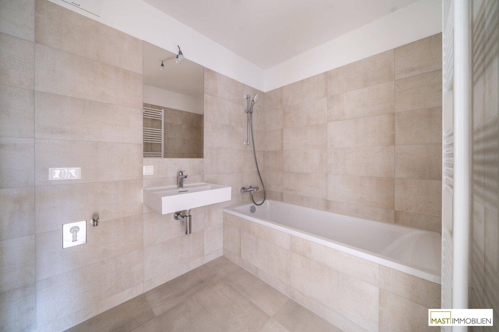 Modernes Badezimmer mit beigefarbenen Fliesen, Badewanne, Waschtisch und beheiztem Handtuchhalter.