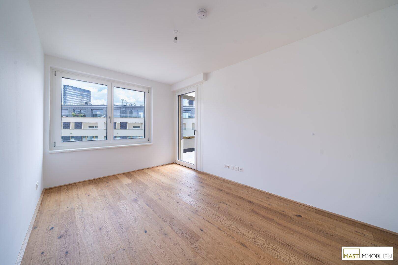 Helles Zimmer mit Holzboden, großen Fenstern und Zugang zu einem Außenbereich.