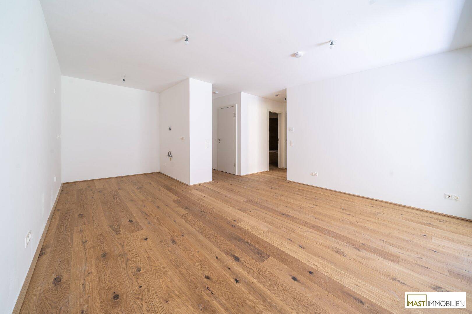 Geräumiger, unmöblierter Wohnbereich mit hellem Holzparkett und weißen Wänden in einem Neubau.