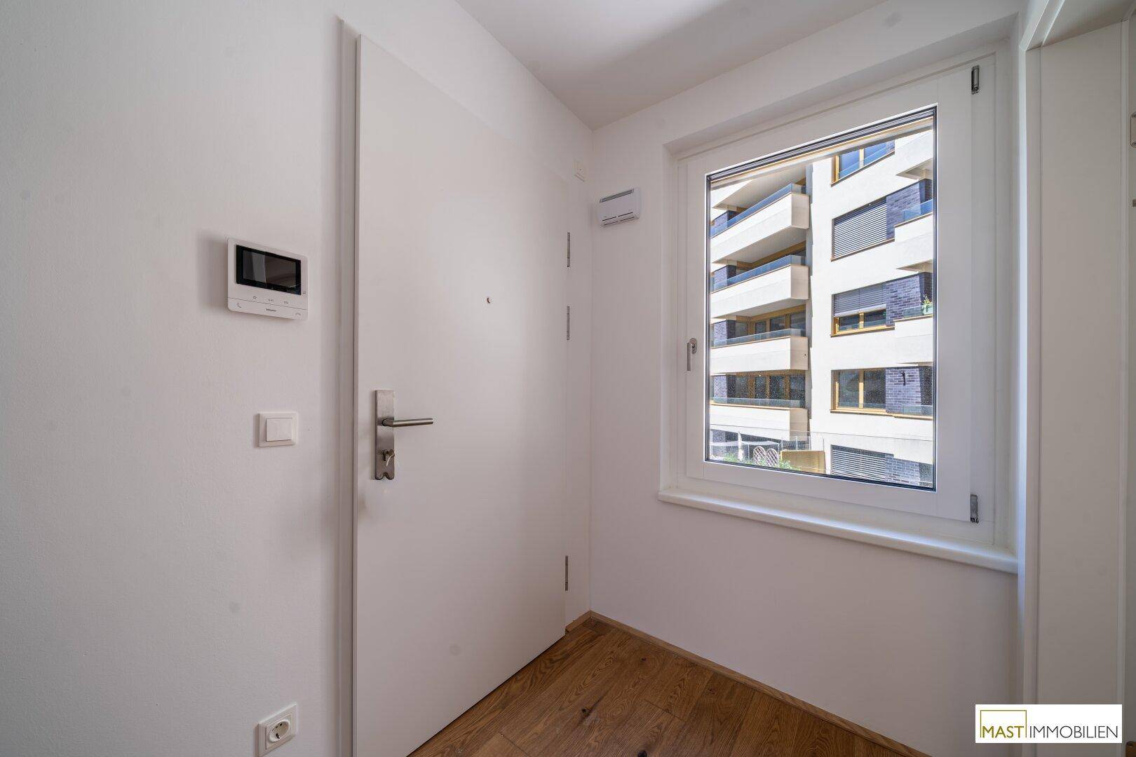 Eingangsbereich mit weißer Tür, Gegensprechanlage und Fenster mit Blick auf die moderne Fassade eines Nachbargebäudes.
