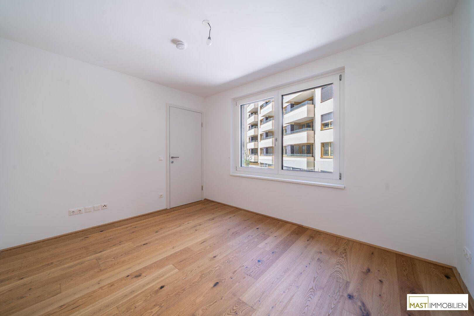 Helles Schlafzimmer mit Parkettboden und großem Fenster, das viel Tageslicht hereinlässt.