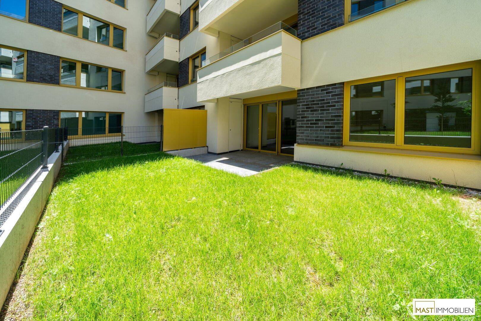 Grüner Gartenbereich mit Rasenfläche und einer kleinen gepflasterten Terrasse vor einem modernen Gebäude.
