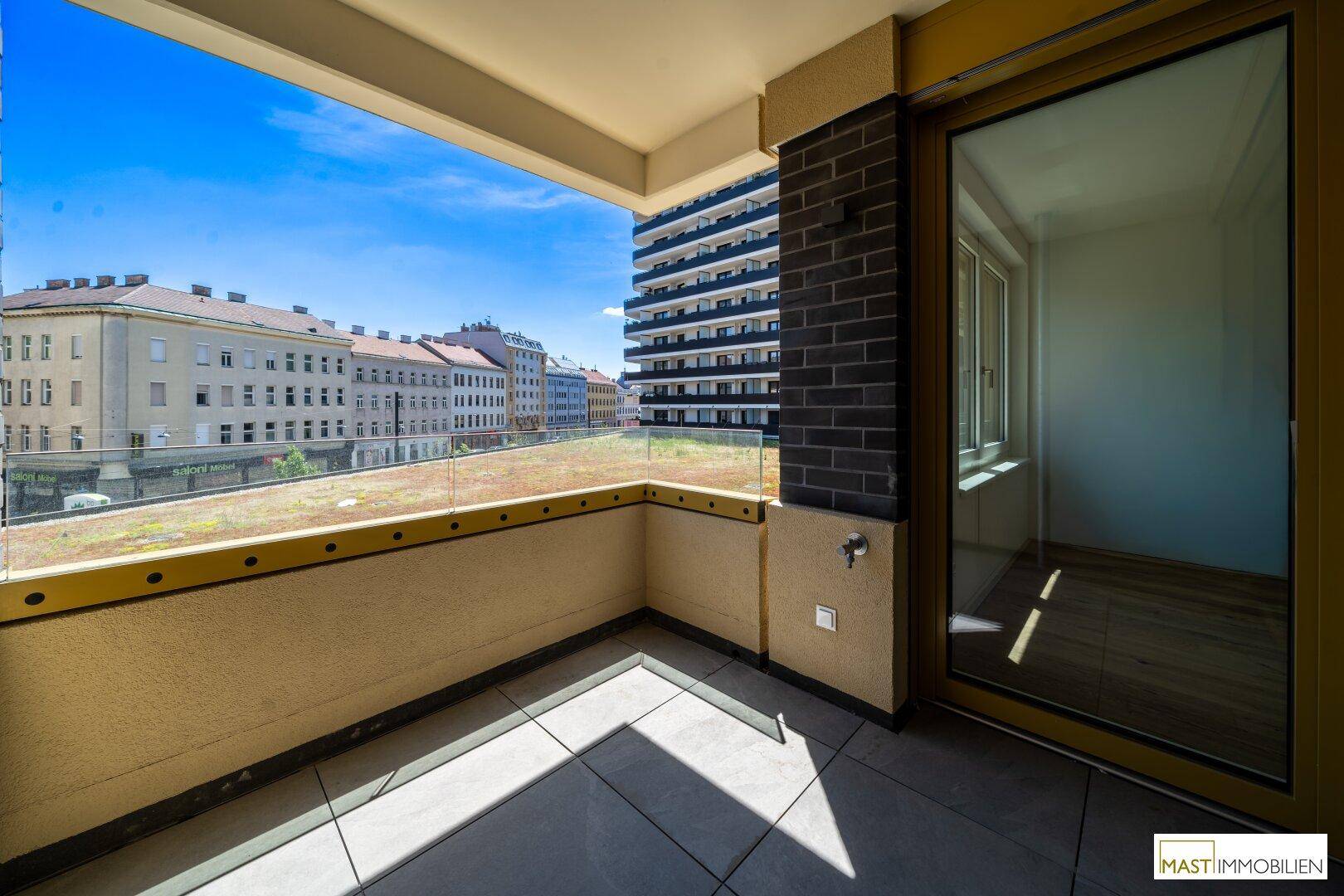 Geräumiger Balkon mit Fliesenboden, Glasgeländer und Blick auf die Stadt sowie ein begrüntes Dach.