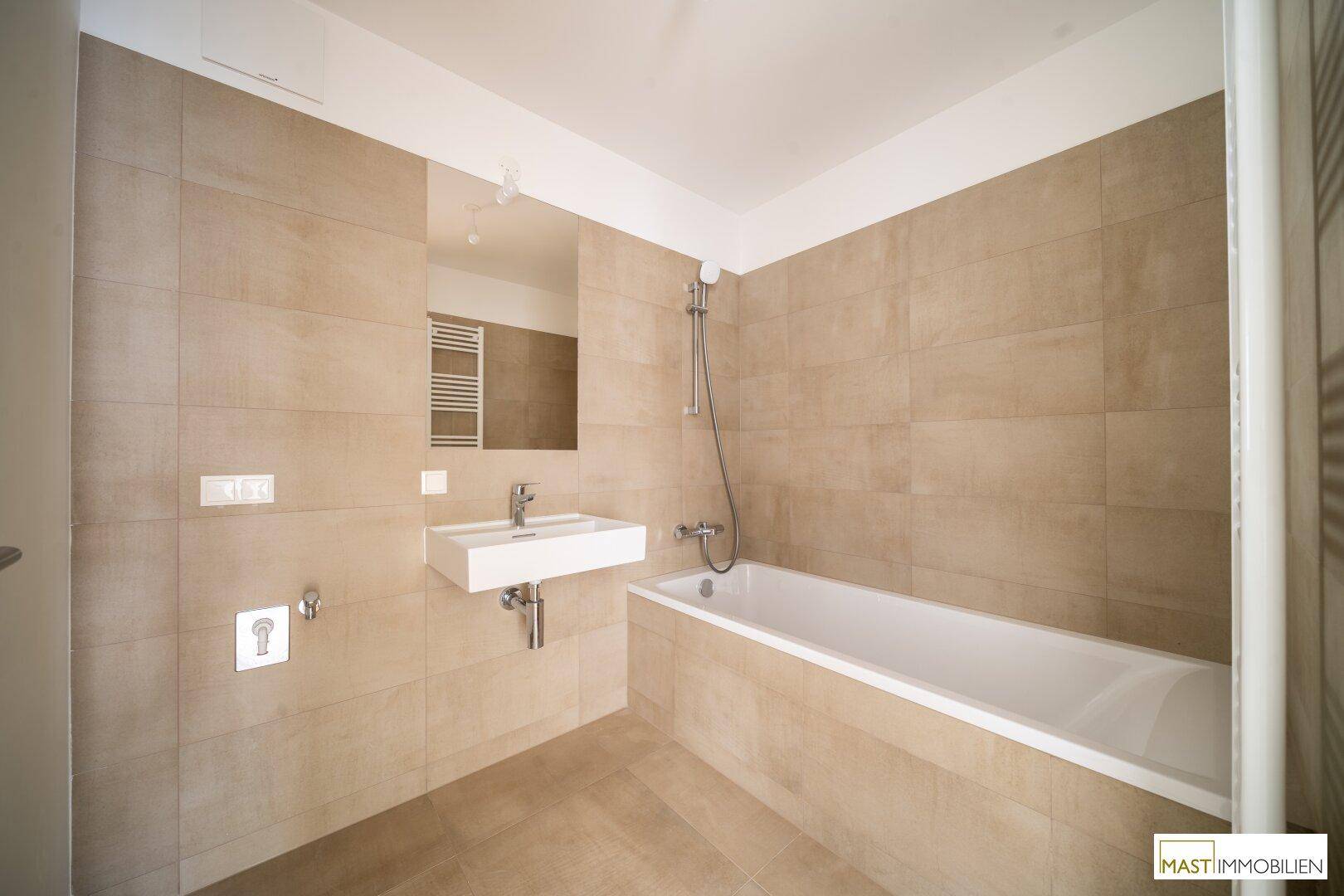 Helles Badezimmer mit beigefarbenen Fliesen, Badewanne mit Duschfunktion und beheiztem Handtuchhalter.
