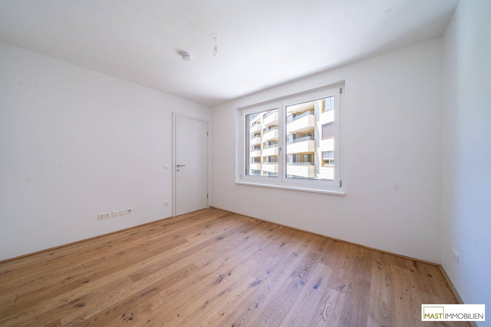 Helles Schlafzimmer mit Parkettboden und Fenster, das auf ein gegenüberliegendes Gebäude blickt.