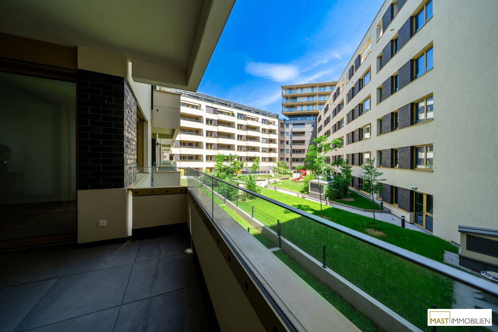 Balkon mit Glasgeländer und Blick auf einen gepflegten grünen Innenhof und moderne Gebäude.