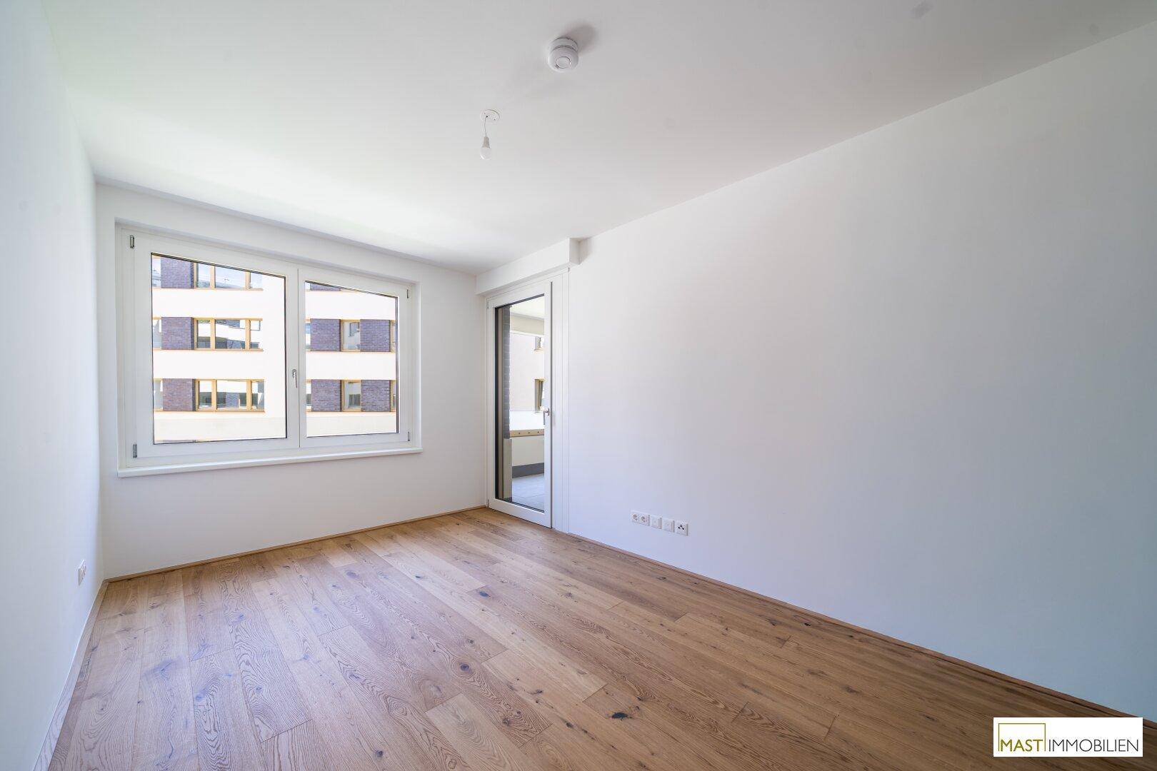 Helles Zimmer mit großem Fenster und Holzboden, bereit für die Einrichtung als Schlaf- oder Arbeitszimmer.