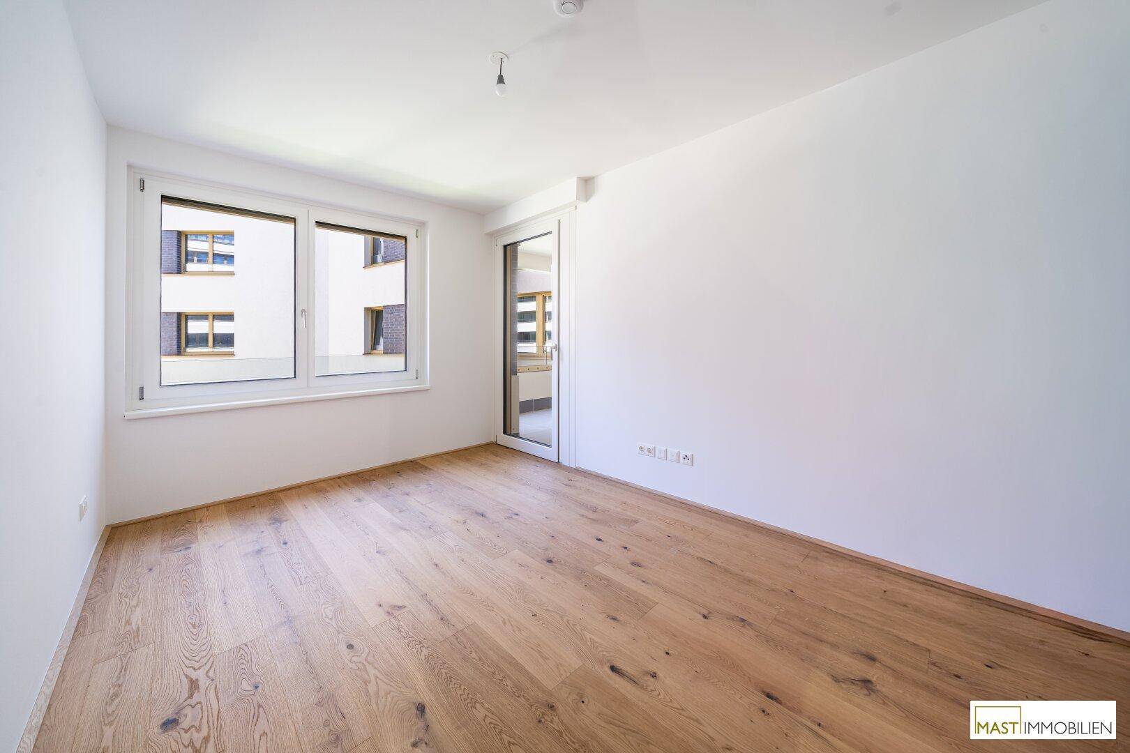 Helles Schlafzimmer mit Parkettboden und Fenster, das viel Tageslicht hereinlässt und einen Balkonzugang bietet.