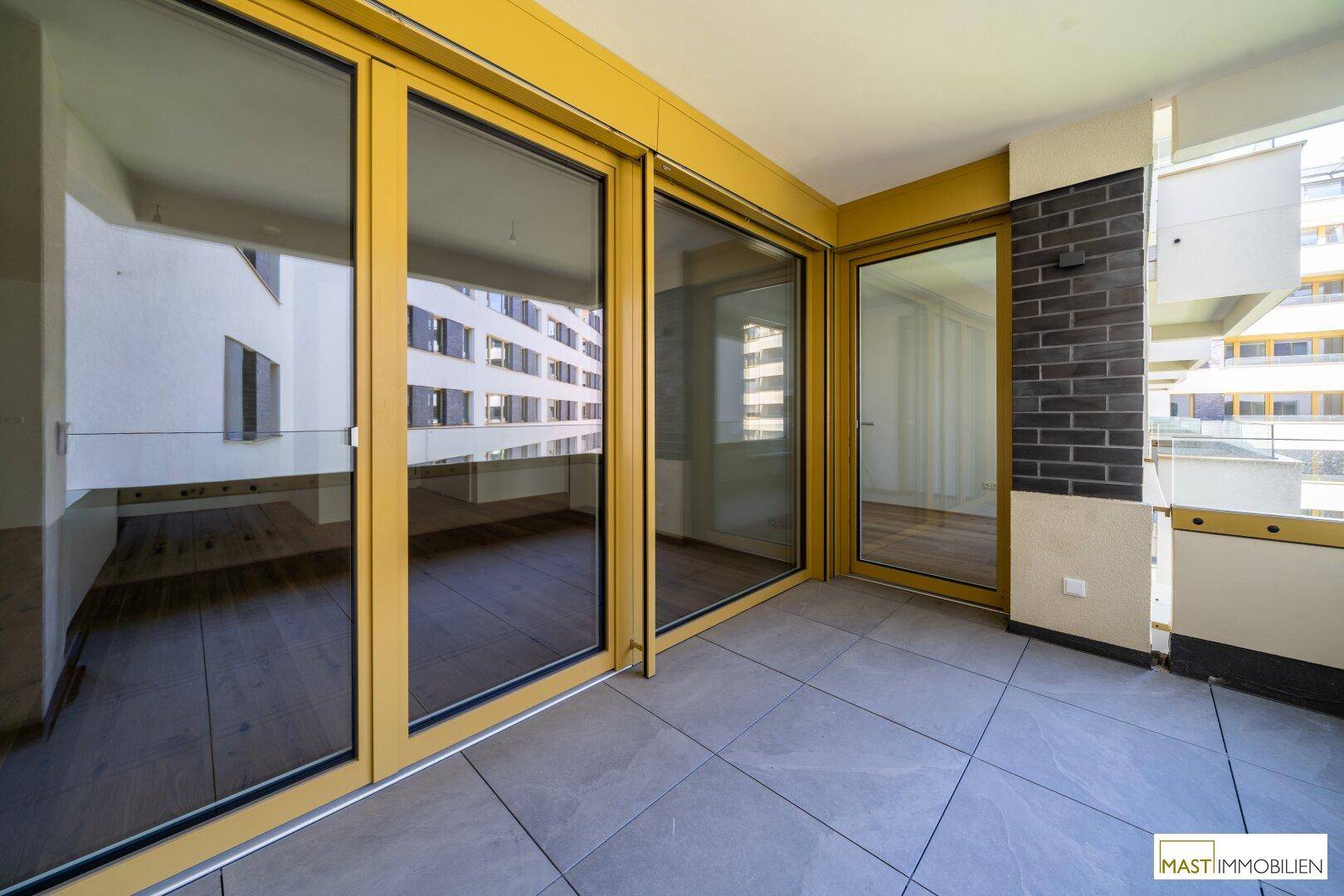 Geräumiger Balkon mit Fliesenboden und gelben Fensterrahmen, der einen Blick auf die Umgebung bietet.