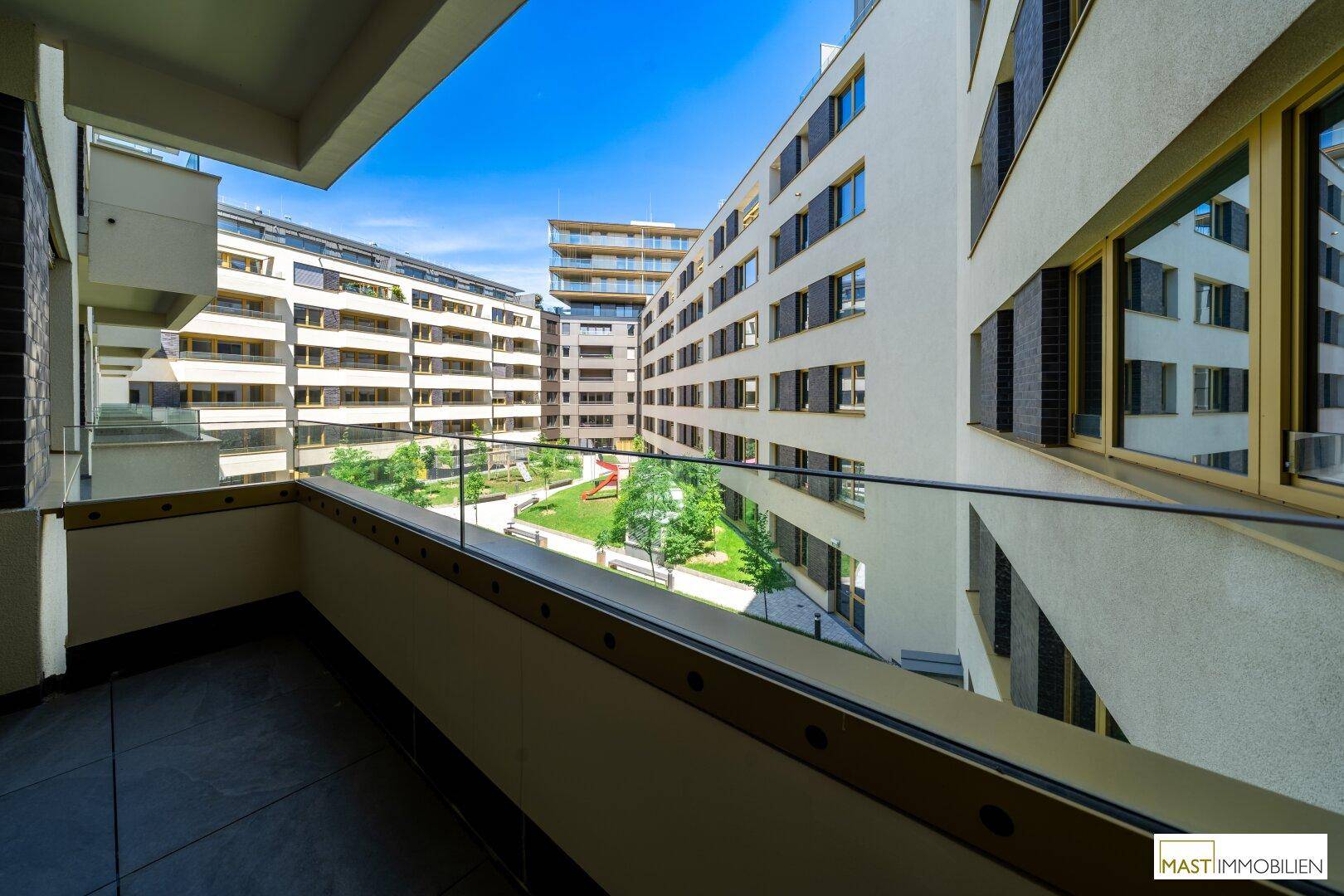 Balkon mit Blick auf einen grünen Innenhof und Spielplatz, umgeben von moderner Wohnbebauung.