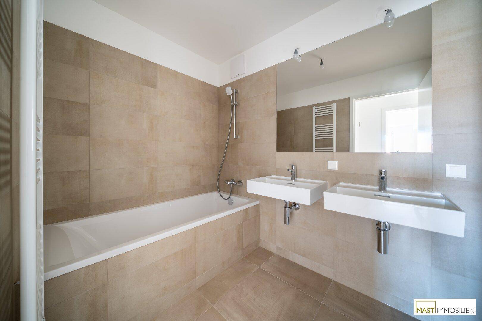 Modernes Badezimmer mit Badewanne, Doppelwaschbecken und großen beigefarbenen Wandfliesen.