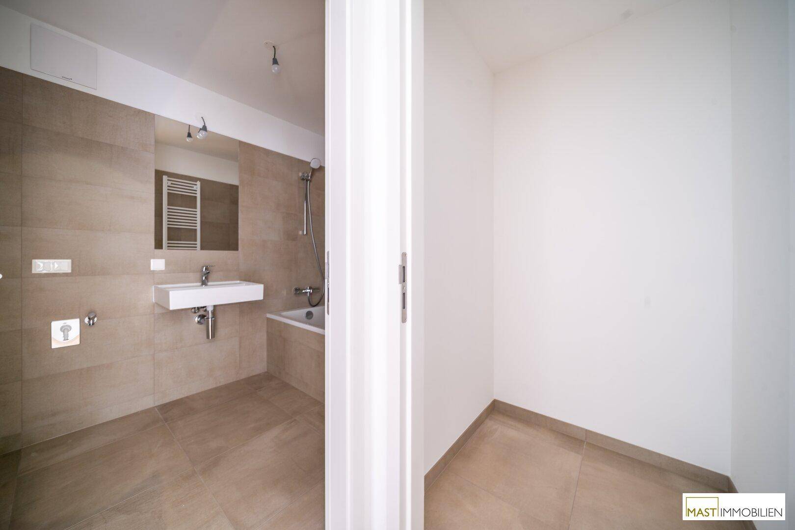 Blick in ein modernes Badezimmer mit beigen Fliesen und angrenzendem leeren Raum.
