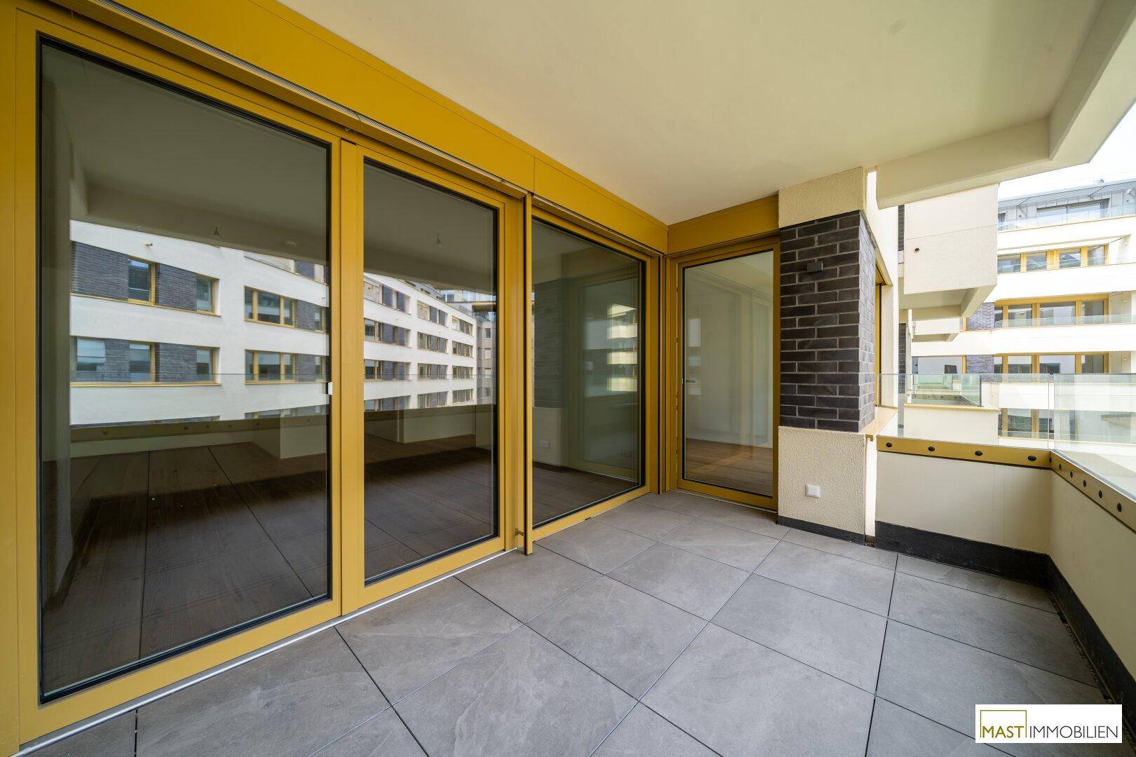 Geräumiger Balkon mit grauen Fliesen und gelben Fensterrahmen, mit Blick auf umliegende Gebäude.