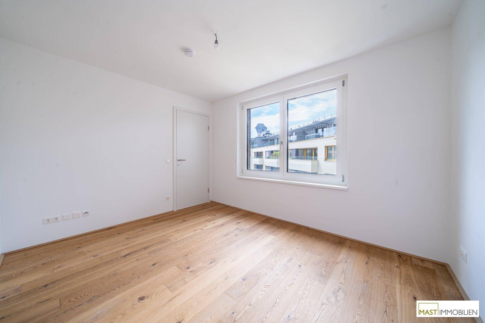 Helles Schlafzimmer mit Holzboden und großem Fenster, bereit zur individuellen Gestaltung.