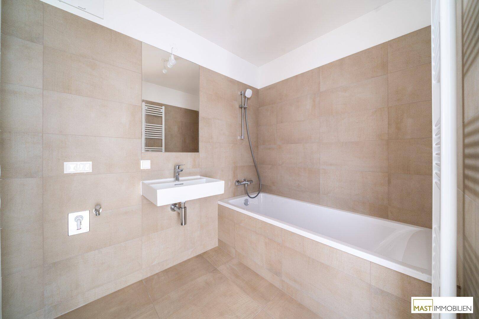 Helles Badezimmer mit beigefarbenen Fliesen, Badewanne mit Duschfunktion und Handtuchheizkörper.
