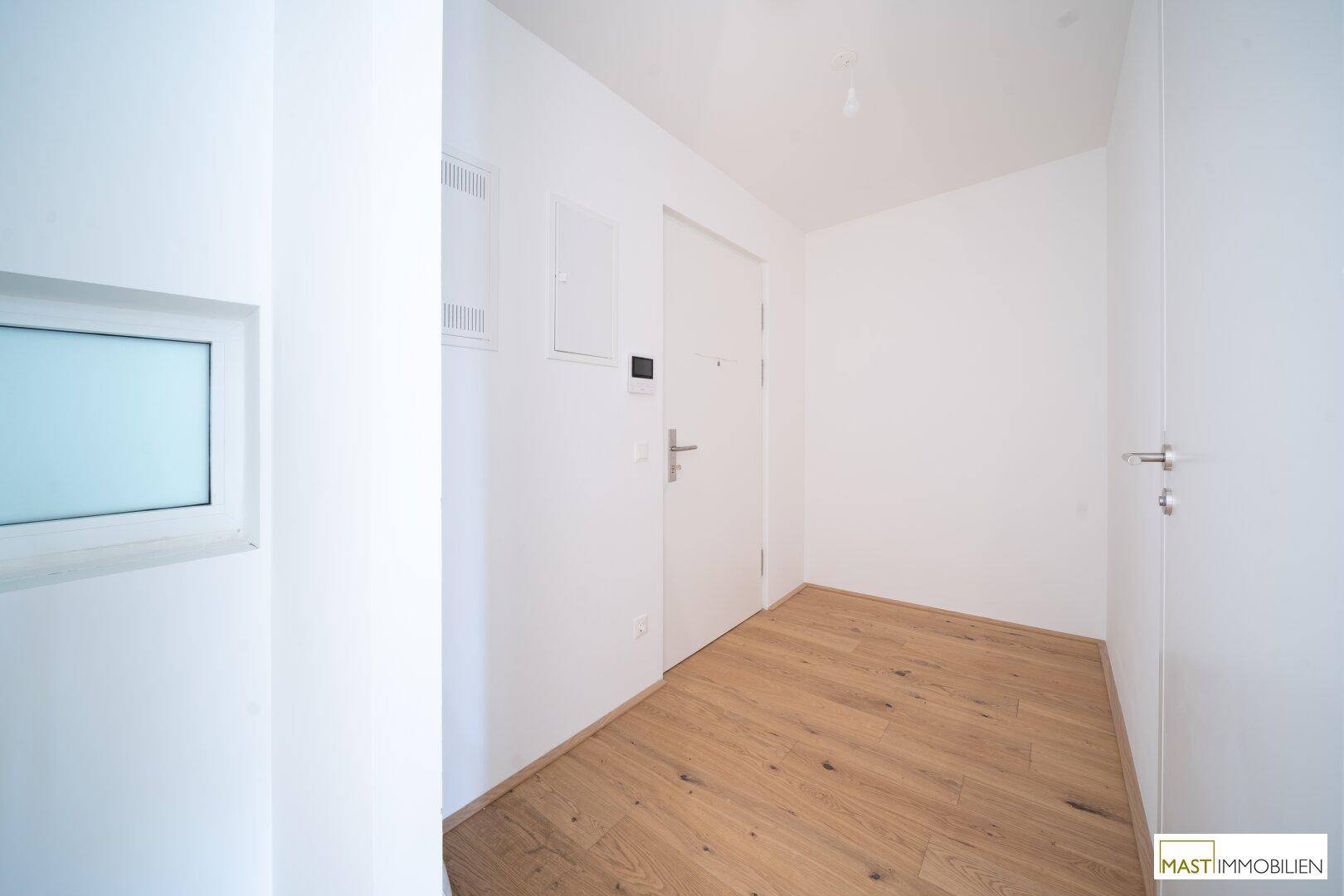 Heller Eingangsbereich mit Holzboden und weiß gestrichenen Wänden, der zu weiteren Räumen führt.