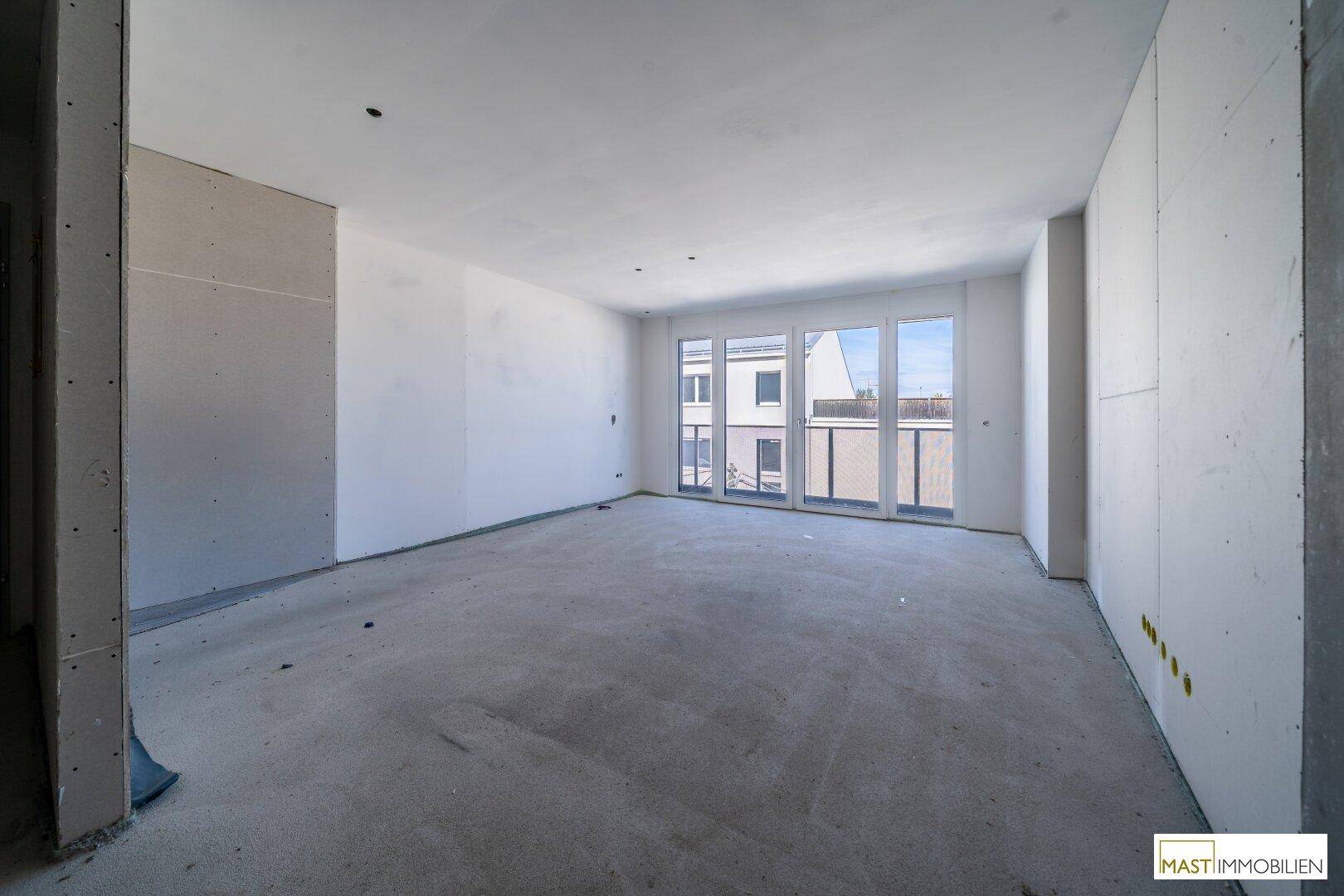 Geräumiger, unfertiger Innenraum mit großen Fenstern und Zugang zu einem potenziellen Balkon.