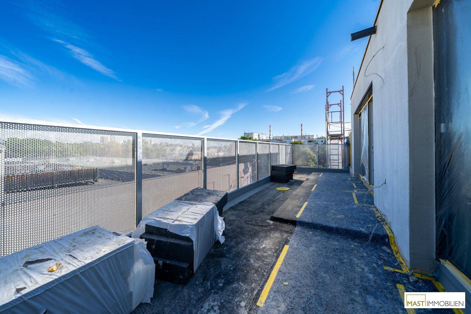 Weitläufige Terrasse eines Neubaus im Rohbau mit Baumaterialien und Gerüst unter blauem Himmel.