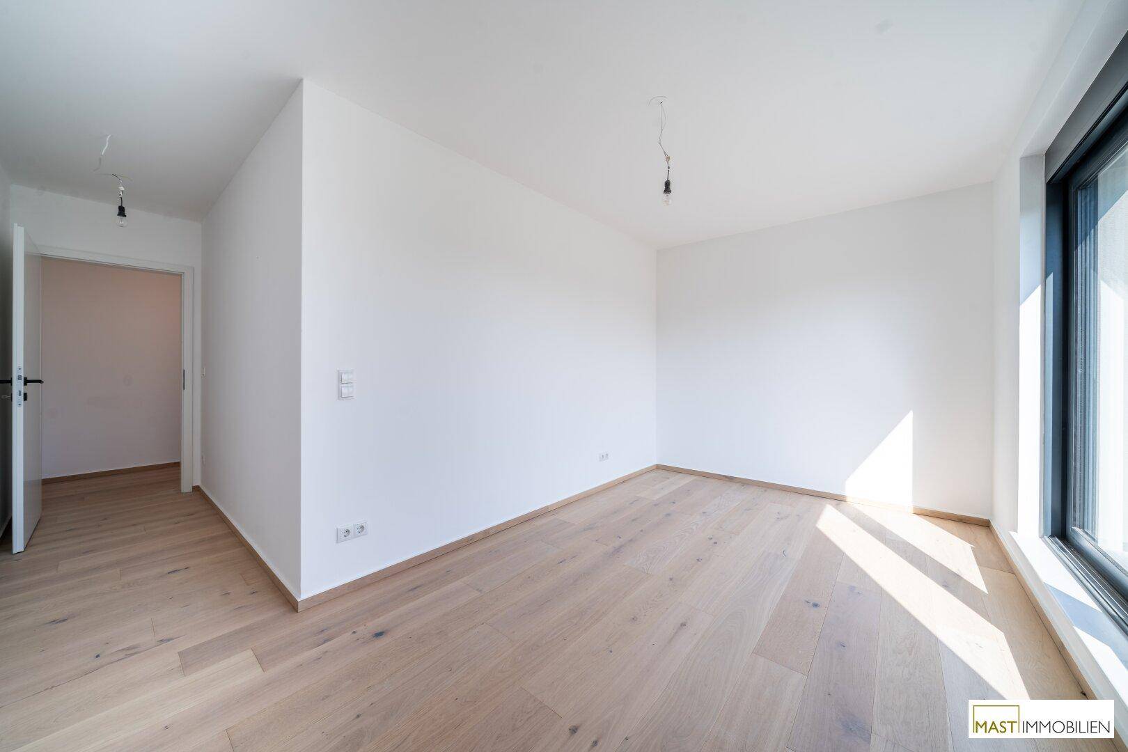 Geräumiges, helles Zimmer mit Holzboden und großem Fenster, das viel Tageslicht hereinlässt.