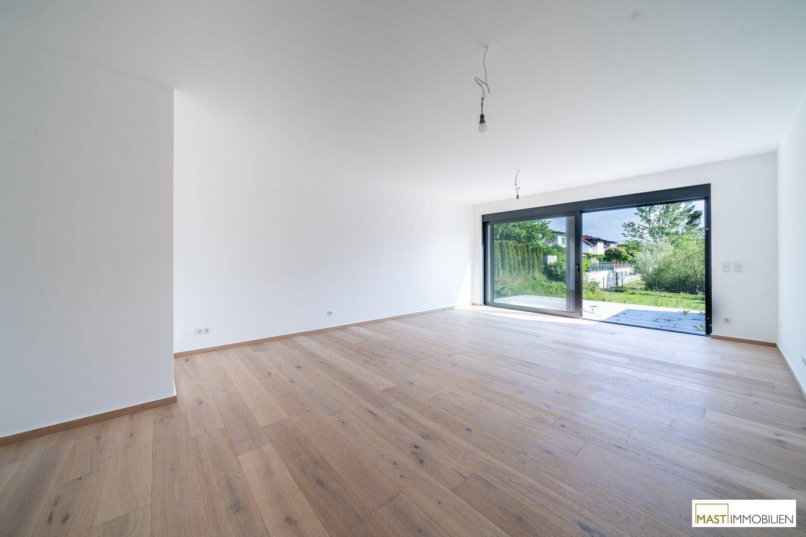 Helles, leeres Zimmer mit Holzboden und großem Fenster mit Zugang zum Garten.