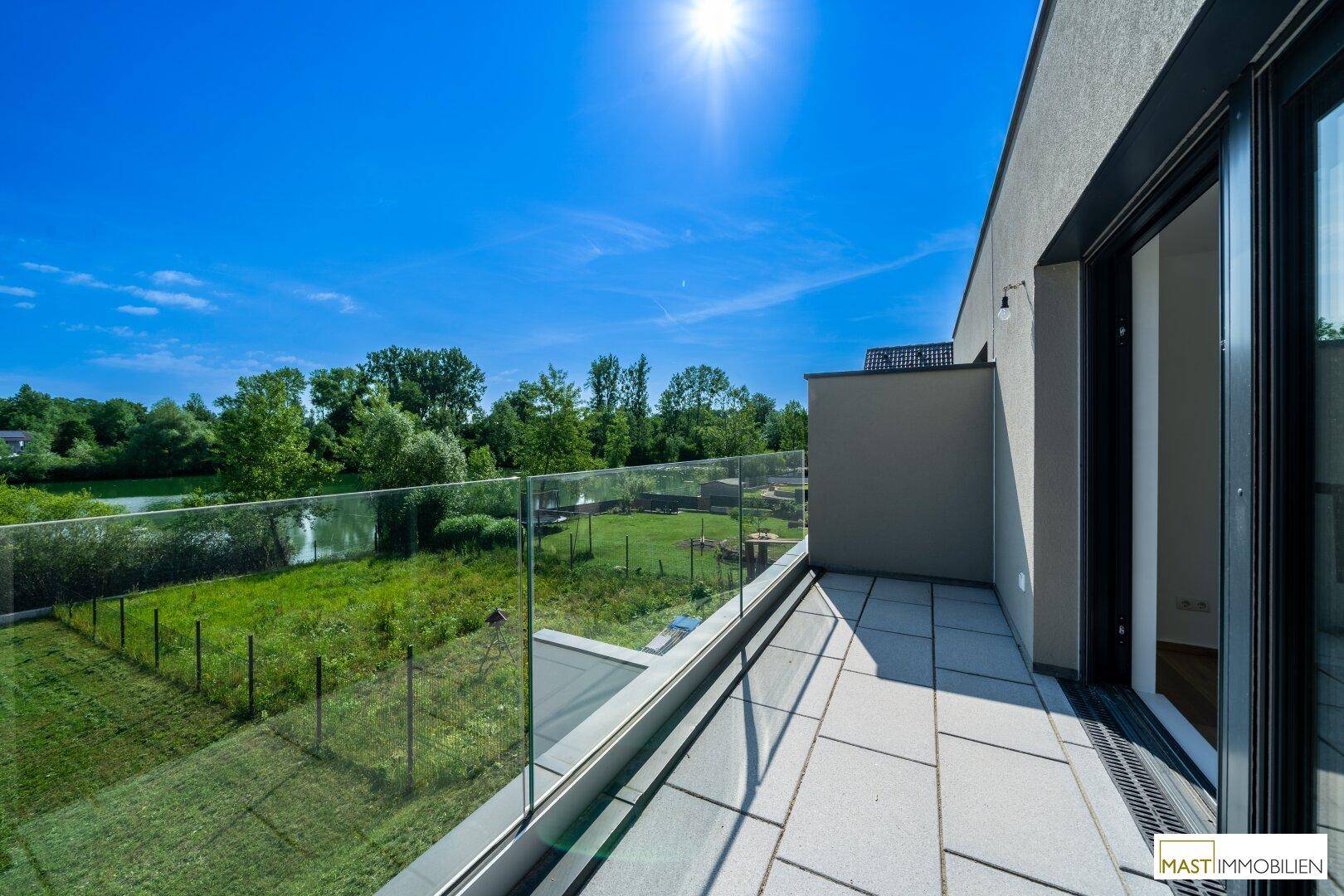 Sonniger Balkon mit Glasgeländer und weitem Blick über grüne Felder und Bäume.